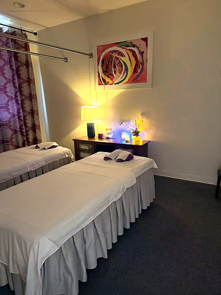The couples room lit by one lamp and candles: two massage tables side by side in white linen with folded towels, under an abstract canvas