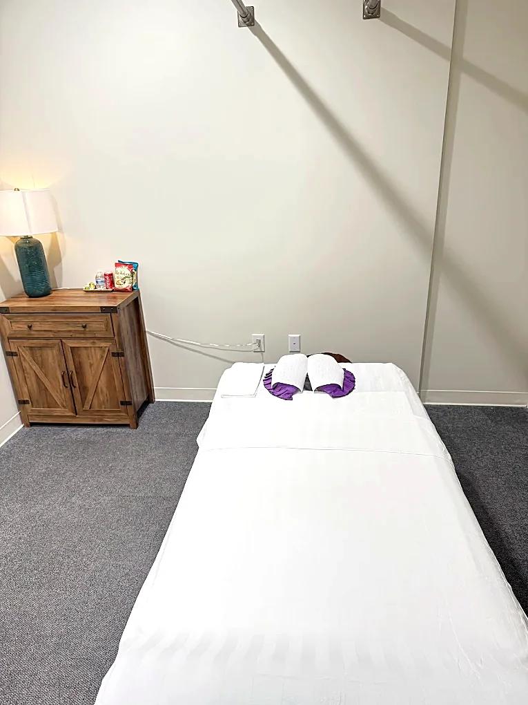 A treatment room with the table made up in white linen, folded towels on a purple wrap, a lamp on a wooden cabinet and bottled water set out