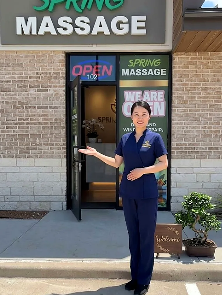 A therapist in navy uniform holding the studio door open beside a wooden Welcome sign and a potted bonsai, under the SPRING MASSAGE sign
